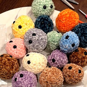 May include: A selection of colourful, plush, round crocheted items with black button accents. The items are in various colours, including yellow, orange, blue, pink, and brown. The items are arranged on a white surface.