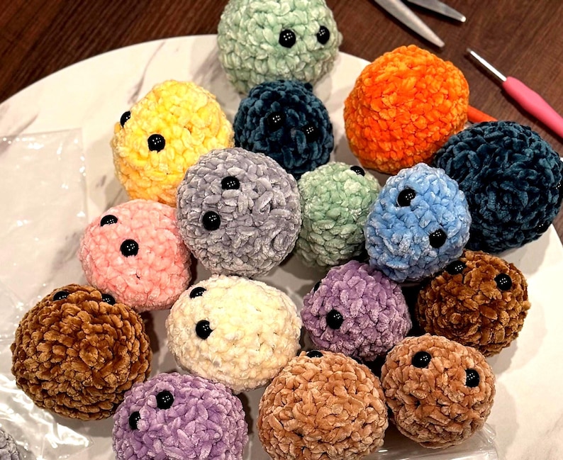 May include: A selection of colourful, plush, round crocheted items with black button accents. The items are in various colours, including yellow, orange, blue, pink, and brown. The items are arranged on a white surface.