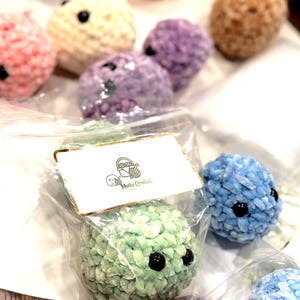 May include: A selection of colourful, crocheted plush toys in shades of pink, purple, and blue. Each plush has black button eyes. One is packaged in a clear bag with a tag that reads "Mochi Crochet".