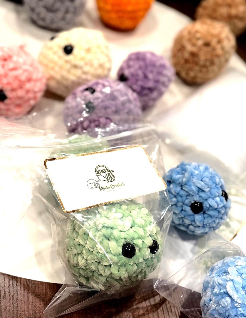 May include: A selection of colourful, crocheted plush toys in shades of pink, purple, and blue. Each plush has black button eyes. One is packaged in a clear bag with a tag that reads "Mochi Crochet".