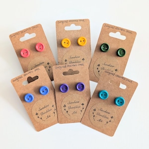 May include: Six pairs of button-shaped stud earrings in various colours including red, yellow, dark green, blue, purple, and teal. Each pair is mounted on a brown cardboard display card labelled Surgical stainless steel and Sunshine Dandelion Art.