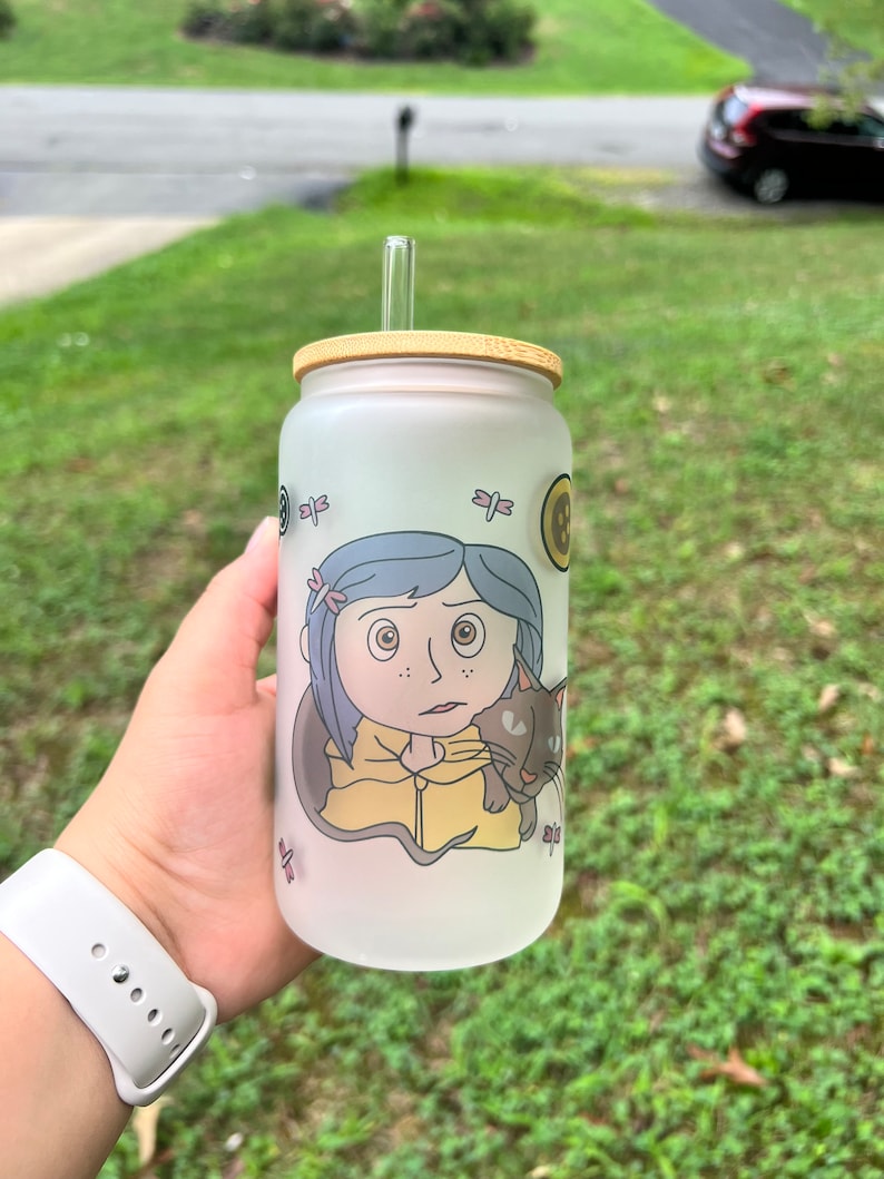 May include: A frosted glass water bottle with a bamboo lid and a straw. The bottle features a cartoon image of a girl with a cat. The girl has blue hair and is wearing a yellow jacket. The cat is black and white.