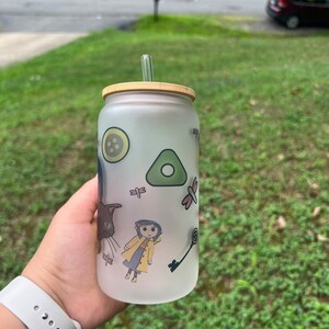 May include: A frosted glass water bottle with a bamboo lid and a straw. The bottle has a whimsical design featuring a girl in a yellow raincoat, a cat, a key, a dragonfly, and a button.