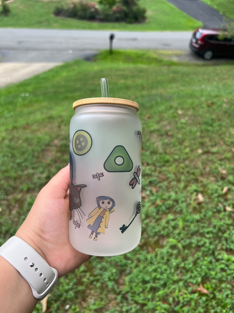 May include: A frosted glass water bottle with a bamboo lid and a straw. The bottle has a whimsical design featuring a girl in a yellow raincoat, a cat, a key, a dragonfly, and a button.