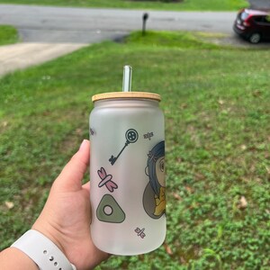 May include: A frosted glass water bottle with a bamboo lid and a clear drinking straw. The bottle has a cartoon character design with a key, a dragonfly, and a green triangle.