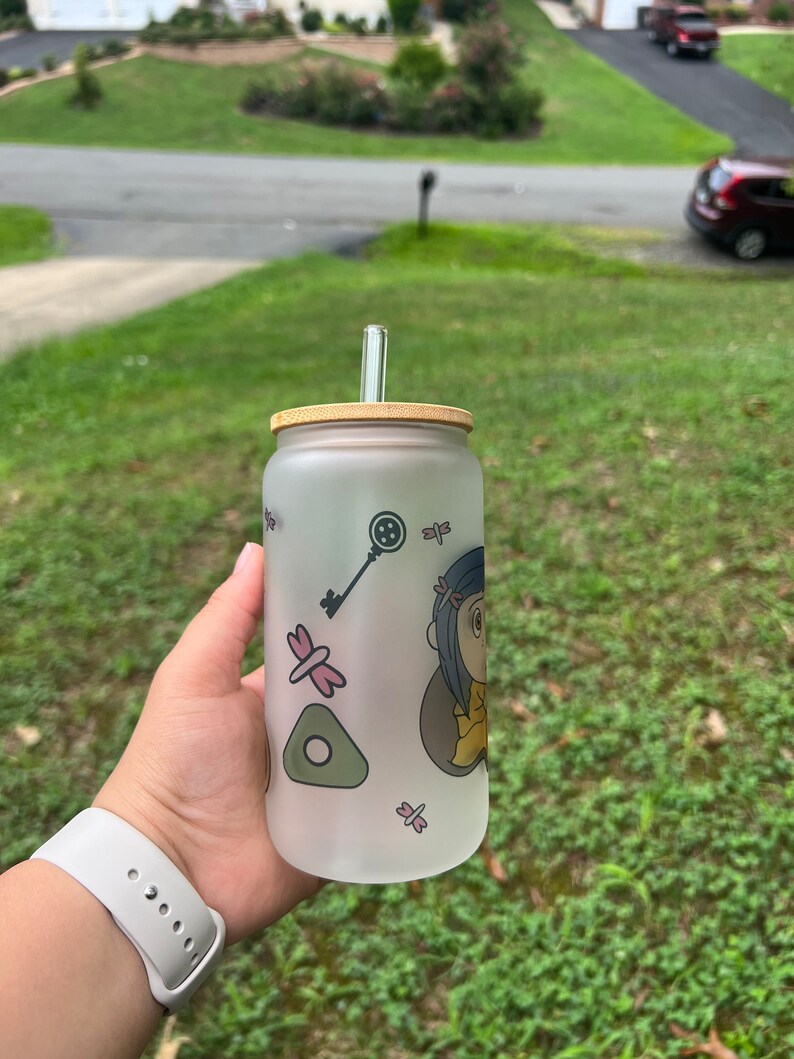 May include: A frosted glass water bottle with a bamboo lid and a clear drinking straw. The bottle has a cartoon character design with a key, a dragonfly, and a green triangle.