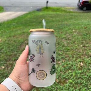 May include: A frosted glass water bottle with a bamboo lid and a glass straw. The bottle has a cartoon character and other designs printed on it.