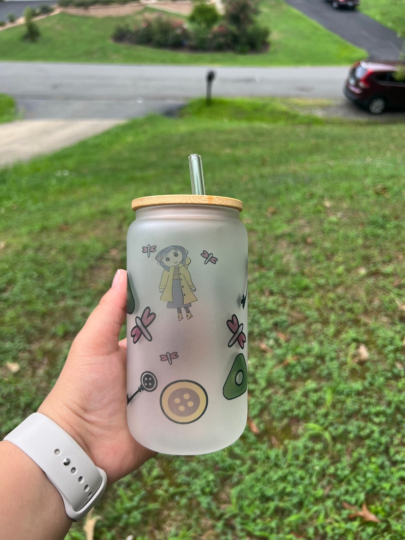 May include: A frosted glass water bottle with a bamboo lid and a glass straw. The bottle has a cartoon character and other designs printed on it.
