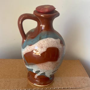 May include: A ceramic pitcher with a handle and a lid. The pitcher is primarily brown with blue and white accents. The lid is red. The pitcher is sitting on a cardboard surface.