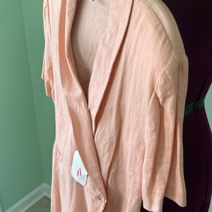 May include: A peach-colored linen wrap dress with a collar and short sleeves. The dress has a tie at the waist and a button closure. The label reads "Adrianna Papell".