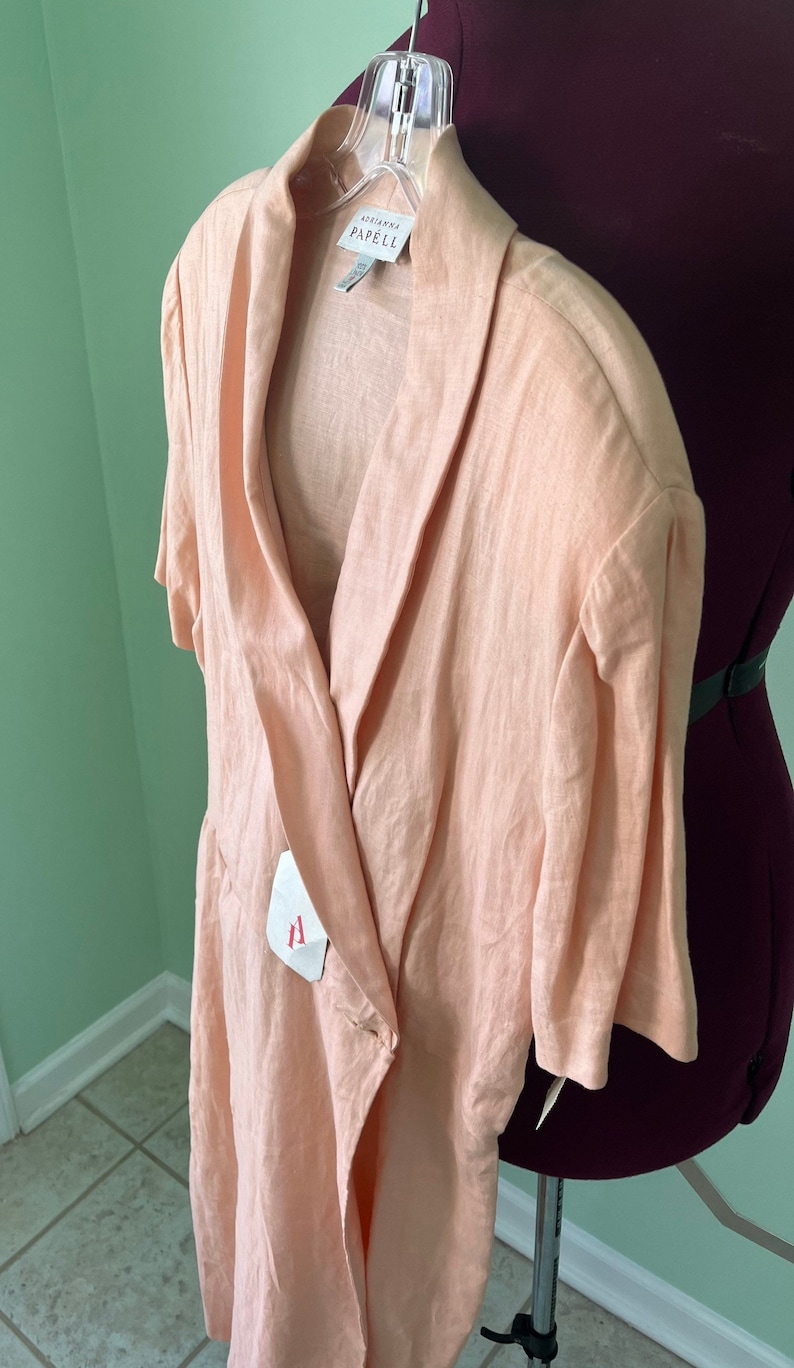 May include: A peach-colored linen wrap dress with a collar and short sleeves. The dress has a tie at the waist and a button closure. The label reads "Adrianna Papell".