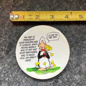 May include: A round, white button with a cartoon penguin holding yellow flowers. Text includes "I love you for that" and a quote about friendship. A yellow measuring tape is above the button, showing 2 inches.