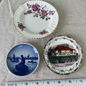 May include: Three decorative ceramic plates. The first plate is white with a pink floral design. The second plate is blue with a mermaid and a sailboat. The third plate is white with a black and gold design and a picture of a shop with the text "The Old Curiosity Shop by Charles Dickens".