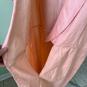 May include: A peach-colored linen dress with a wrap-style front and a button closure. The dress is open in the front, revealing the lining.