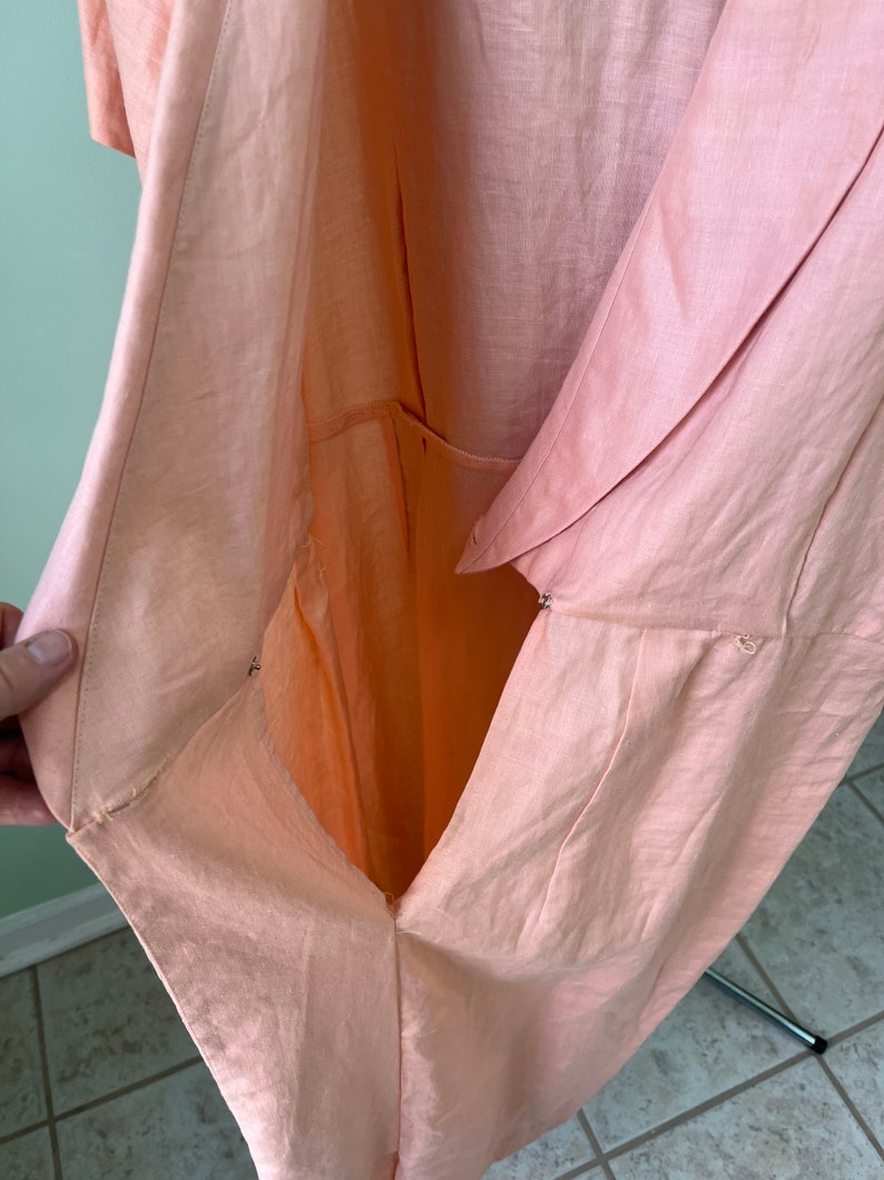 May include: A peach-colored linen dress with a wrap-style front and a button closure. The dress is open in the front, revealing the lining.