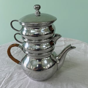 May include: A silver three-tiered coffee pot with a wooden handle. The pot has a lid and a spout.