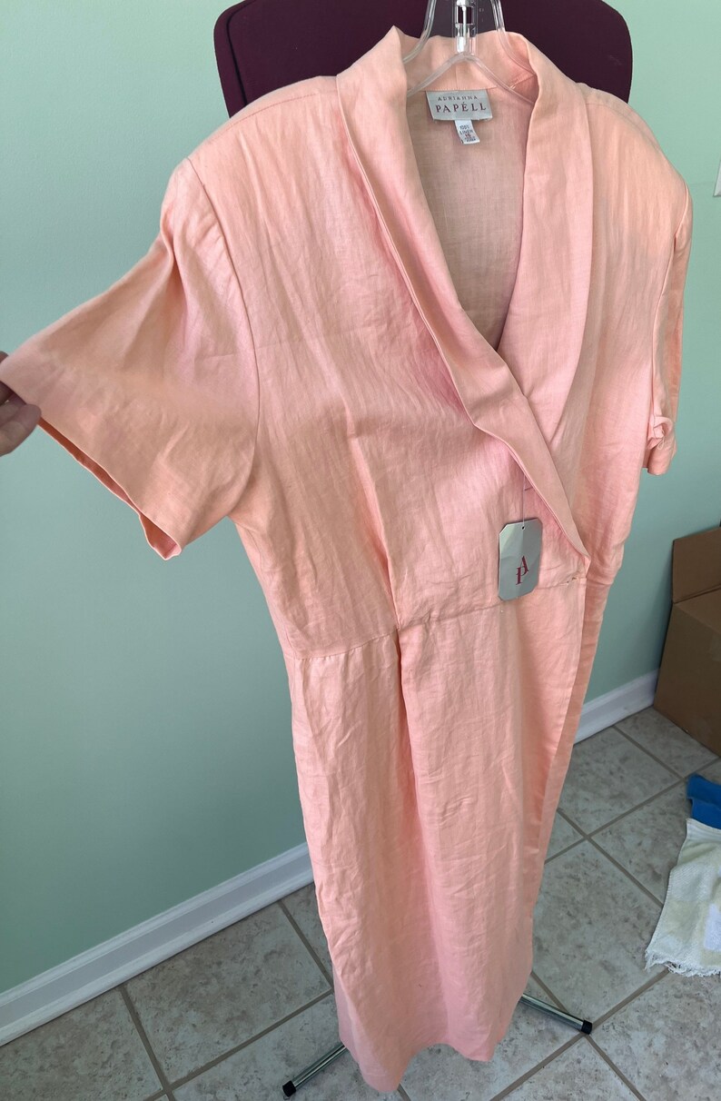 May include: A peach-colored linen dress with a wrap-style neckline and short sleeves. The dress has a tag that reads "Adriana Papell".