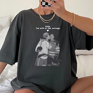 May include: Dark grey t-shirt with a black and white image of two people embracing. The text "help, I'm still at the cottage" is above the image. The shirt also has the text "COMFORT COLORS PEPPER" and "design available in multiple colors!"