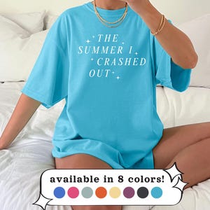 Pode incluir: Camiseta azul lagoa com o texto "THE SUMMER I CRASHED OUT" em branco. A camiseta faz parte da cole&ccedil;&atilde;o "COMFORT COLORS" e est&aacute; dispon&iacute;vel em 8 cores. A modelo est&aacute; usando joias douradas.