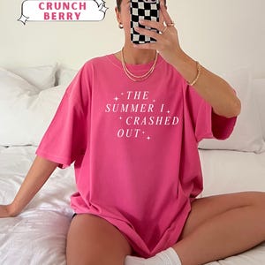 Pode incluir: Uma t-shirt rosa brilhante com o texto branco "THE SUMMER I CRASHED OUT". A t-shirt &eacute; rotulada como "Crunch Berry" da cole&ccedil;&atilde;o "Comfort Colors". A pessoa est&aacute; usando joias de ouro e meias brancas.