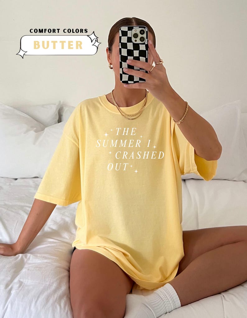 Pode incluir: Camiseta amarela clara com o texto branco "THE SUMMER I CRASHED OUT". A camisa est&aacute; rotulada como "BUTTER" em "COMFORT COLORS". A modelo est&aacute; usando joias douradas e segurando um telefone com uma capa xadrez em preto e branco.