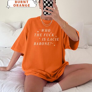May include: Burnt orange t-shirt with the text "WHO THE FUCK IS LACIE BARONE?" in white. The shirt is a comfort color and has short sleeves. The image also includes the text "COMFORT COLORS BURNT ORANGE".