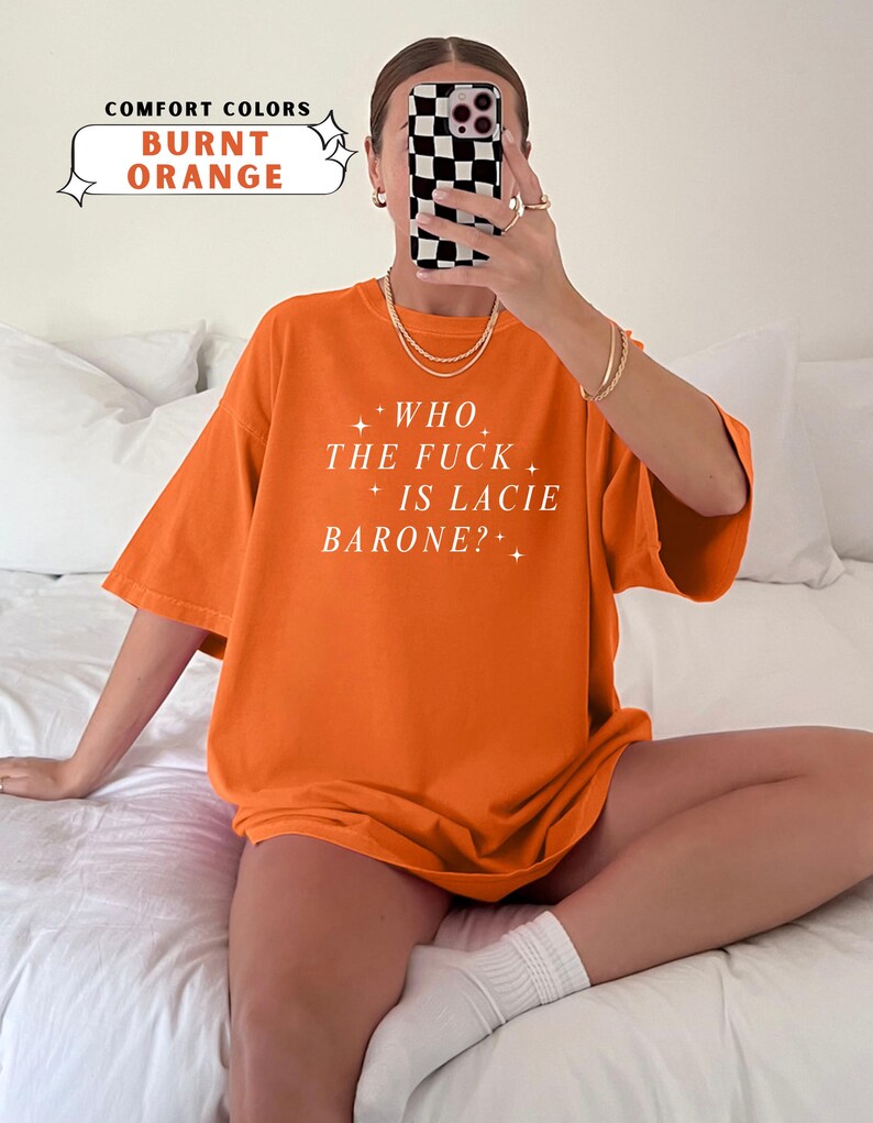 May include: Burnt orange t-shirt with the text "WHO THE FUCK IS LACIE BARONE?" in white. The shirt is a comfort color and has short sleeves. The image also includes the text "COMFORT COLORS BURNT ORANGE".