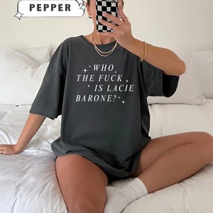 May include: Dark grey t-shirt with the text "WHO THE FUCK IS LACIE BARONE?" in white. The shirt also has the text "COMFORT COLORS" and the color "PEPPER" in a white box. The shirt is worn by a person.