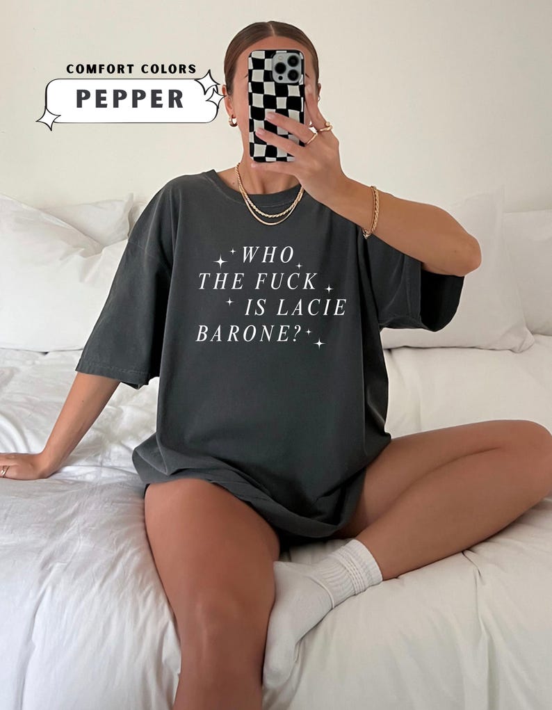 May include: Dark grey t-shirt with the text "WHO THE FUCK IS LACIE BARONE?" in white. The shirt also has the text "COMFORT COLORS" and the color "PEPPER" in a white box. The shirt is worn by a person.