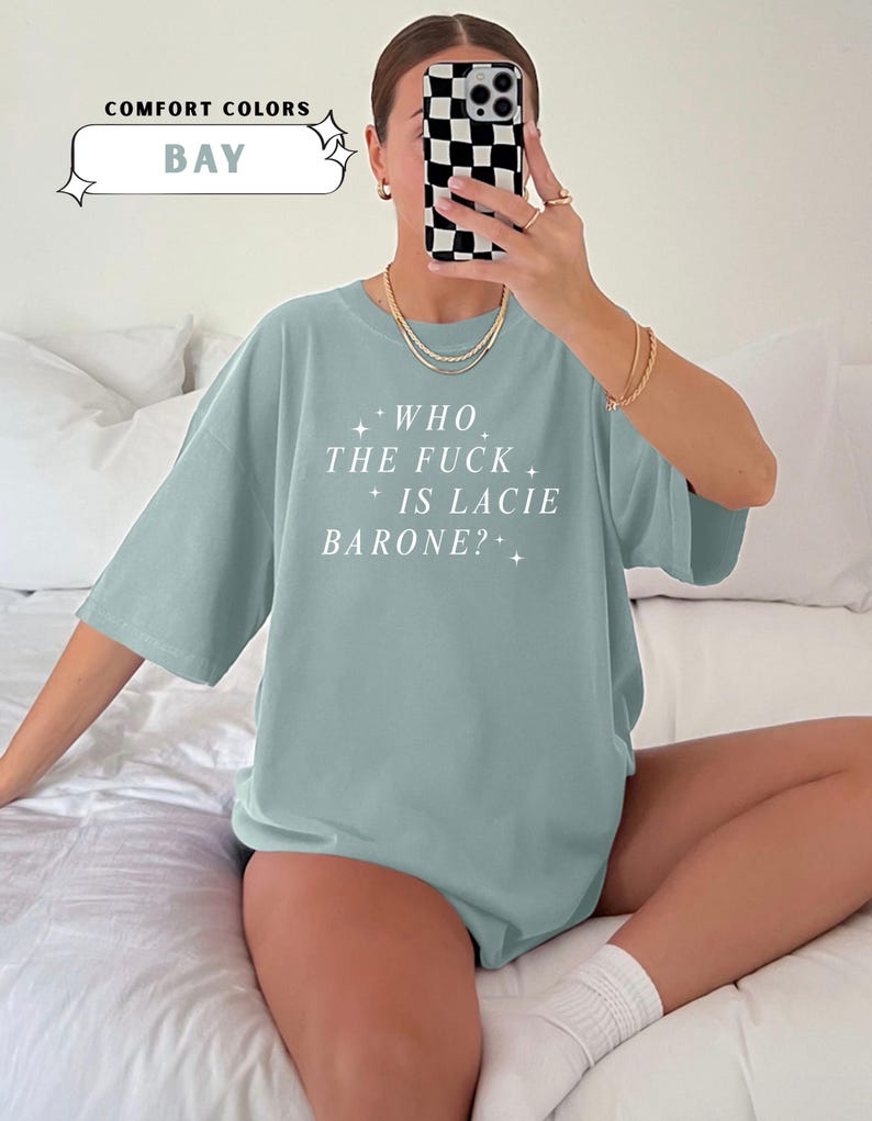May include: A light blue t-shirt with the text "WHO THE FUCK IS LACIE BARONE?" in white. The shirt is part of the "Comfort Colors" collection and is in the color "BAY". The shirt has short sleeves and a round neckline.