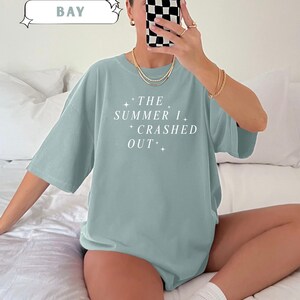 Pode incluir: T-shirt azul claro com o texto branco "THE SUMMER I CRASHED OUT". Faz parte da cole&ccedil;&atilde;o "Comfort Colors" e &eacute; na cor "BAY". Uma pe&ccedil;a de roupa casual.