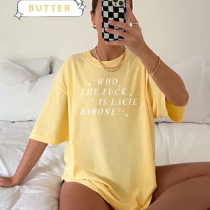 May include: A yellow t-shirt with the text "WHO THE FUCK IS LACIE BARONE?" in white. The shirt is labeled "COMFORT COLORS" and "BUTTER". The model is wearing gold jewelry and holding a phone with a black and white checkered case.