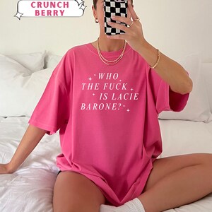 May include: A bright pink t-shirt with the text "WHO THE FUCK IS LACIE BARONE?" in white. The shirt is labeled "Crunch Berry" from the "Comfort Colors" collection. The model is wearing gold jewelry and white socks.