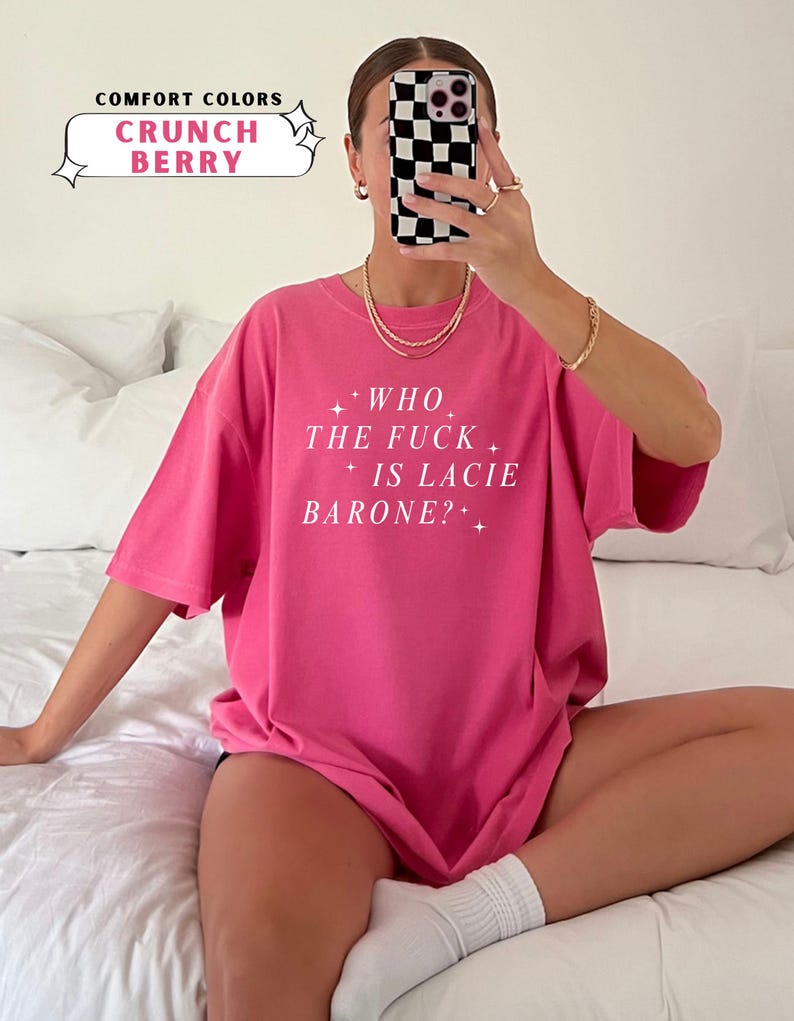 May include: A bright pink t-shirt with the text "WHO THE FUCK IS LACIE BARONE?" in white. The shirt is labeled "Crunch Berry" from the "Comfort Colors" collection. The model is wearing gold jewelry and white socks.