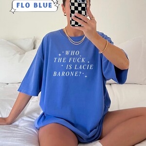 May include: A Flo Blue t-shirt with the text "WHO THE FUCK IS LACIE BARONE?" in white. The shirt is part of the Comfort Colors collection. The image also shows a person holding a phone with a black and white checkered case.