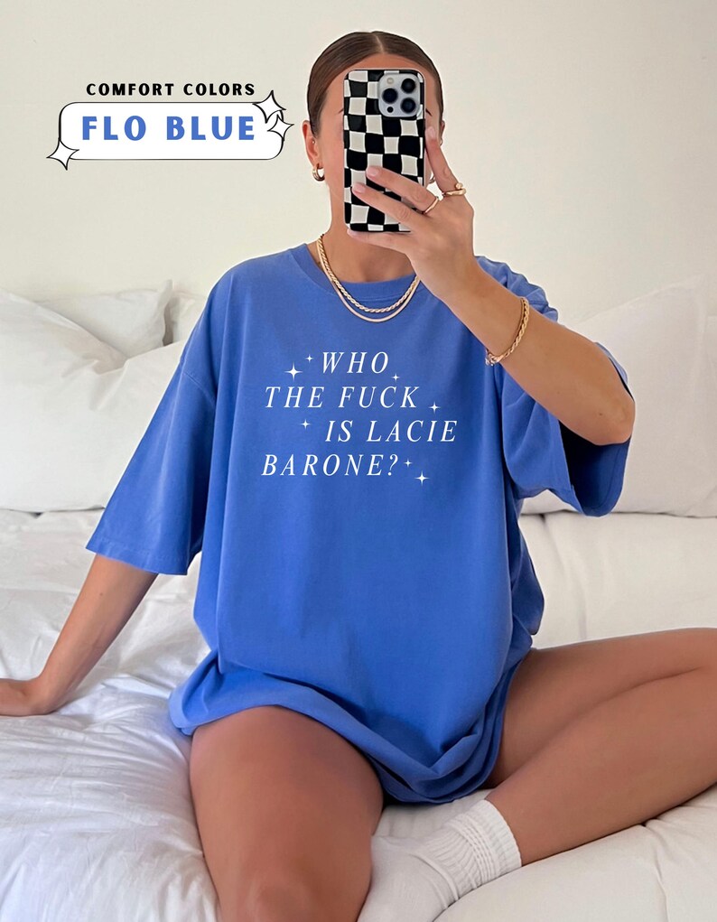 May include: A Flo Blue t-shirt with the text "WHO THE FUCK IS LACIE BARONE?" in white. The shirt is part of the Comfort Colors collection. The image also shows a person holding a phone with a black and white checkered case.