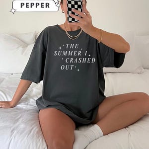 Pode incluir: T-shirt Comfort Colors cinza escuro com o texto branco "THE SUMMER I CRASHED OUT". A camisa tem um ajuste folgado. A palavra "PEPPER" est&aacute; em uma caixa branca no canto superior esquerdo.