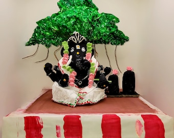 May include: A black Ganesha statue with green and pink flower garlands, sitting on a white and red striped platform under a green paper mache tree.