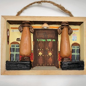 May include: A wooden shadow box featuring a detailed diorama of a traditional building facade. The scene includes two brown pillars, a carved wooden door, and architectural details. The box is framed in natural wood and has a rope for hanging.