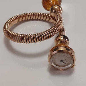 Op de afbeelding: Een goudkleurig spiraalarmbandhorloge met een flexibele, gedraaide metalen band. Het ene uiteinde van de spiraal eindigt in een ronde, barnsteenkleurige glazen kraal, terwijl het andere uiteinde een kleine, ronde wijzerplaat heeft met een witte achtergrond en gouden wijzers.