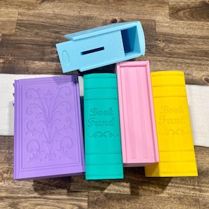 Puede incluir: Una colección de huchas con forma de libro de colores. Las cajas son en tonos morado, turquesa, rosa y amarillo, con la inscripción "Book Fund" en algunas. También hay una caja azul claro con una ranura. Los artículos están dispuestos sobre una superficie de madera.