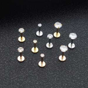 May include: An assortment of lip piercing studs featuring clear, round cubic zirconia stones. The studs come in gold, silver, and rose gold finishes. The jewellery is presented on a dark, textured surface.
