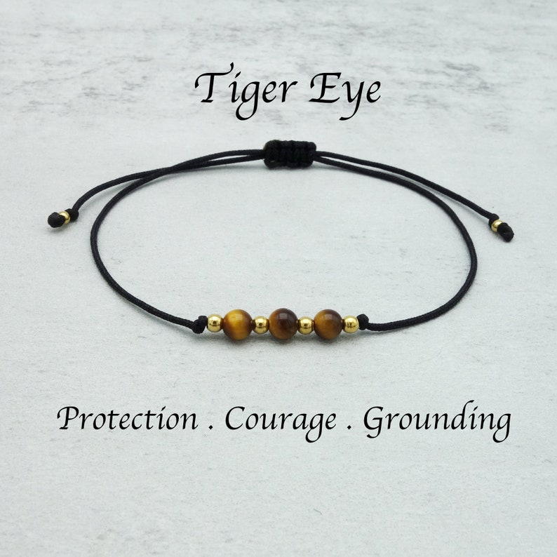 May include: A black cord bracelet with three tiger eye beads and two gold beads. The text "Tiger Eye" is above the bracelet, and the words "Protection . Courage . Grounding" are below.