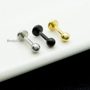 May include: Three labret stud earrings in silver, black, and gold. Each stud has a flat disc on one end and a small ball on the other, joined by a straight post. The earrings are shown on a white surface.