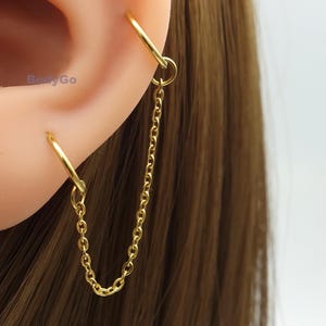 May include: Gold-coloured chain earrings. The earrings feature a small hoop that connects to a chain, which then connects to another hoop. The earrings are designed to be worn through two piercings, with the chain hanging between them.