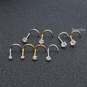 May include: A selection of nose rings in gold and silver hues, each adorned with a clear, sparkling gemstone. The jewellery is displayed on a dark surface, highlighting the different styles and finishes.