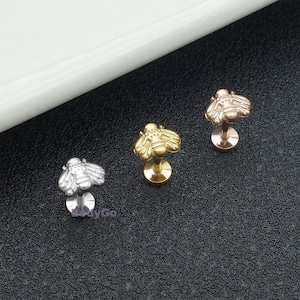 May include: Three bee-shaped lip piercing studs in silver, gold, and rose gold. Each stud features a detailed bee design on a flat disc base. The studs are displayed on a black surface with a white curved edge in the background.