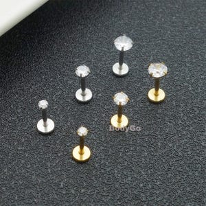 May include: Six labret studs with clear, round, prong-set gemstones. Three studs feature a silver-toned metal post and flat back, while three feature a gold-toned metal post and flat back. The text BodyGo is visible in the lower centre area.