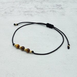 May include: Black cord bracelet with four tiger eye beads and gold-tone details. The bracelet is adjustable with a sliding knot. A minimalist design, suitable for everyday wear. The bracelet is adjustable.