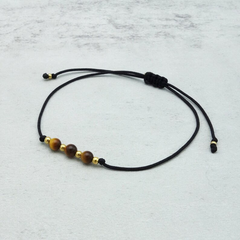 May include: Black cord bracelet with four tiger eye beads and gold-tone details. The bracelet is adjustable with a sliding knot. A minimalist design, suitable for everyday wear. The bracelet is adjustable.